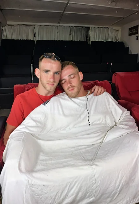 A man sleeping in a movie theater bench seat church pew with a blanket snuggled up next to another male. They are both covered up in a blanket, snuggled together, head on shoulder,