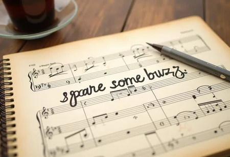 A music sheet, that has some notes written in it. On the center of the sheet there is hand written "spare some buzz" written in beautiful handwritten style with a stylo. There is some coffee in the background
