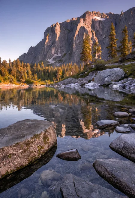 A serene, dreamlike mountain valley at golden hour, the sky awash in soft pastel hues of peach, lavender, and rose gold, long golden sunlight casting dramatic shadows across the landscape. In the foreground, a crystal-clear alpine lake reflects the colors of the sky and surrounding snow-dusted pine trees, with delicate ripples forming soft patterns on the water's surface. Moss-covered rocks line the lake’s edge, and patches of wildflowers in muted tones (soft pinks, light blues, and creamy whites) dot the grassy banks. Towering granite cliffs rise in the background, their rugged surfaces catching the last warm light of day, while a low mist begins to gather near their base, adding depth and atmosphere. The air is still, crisp, and filled with quiet magic. Shot using a full-frame DSLR camera (Canon EOS R5) with a 35mm f/1.4 prime lens for shallow depth of field and cinematic sharpness. The composition uses rule of thirds, with leading lines from the rocks guiding the eye toward the distant cliffs. Hyperrealistic, ultra-detailed, 8K resolution, soft contrast, fine grain texture, perfect natural lighting, no people, no animals — just untouched, tranquil wilderness.