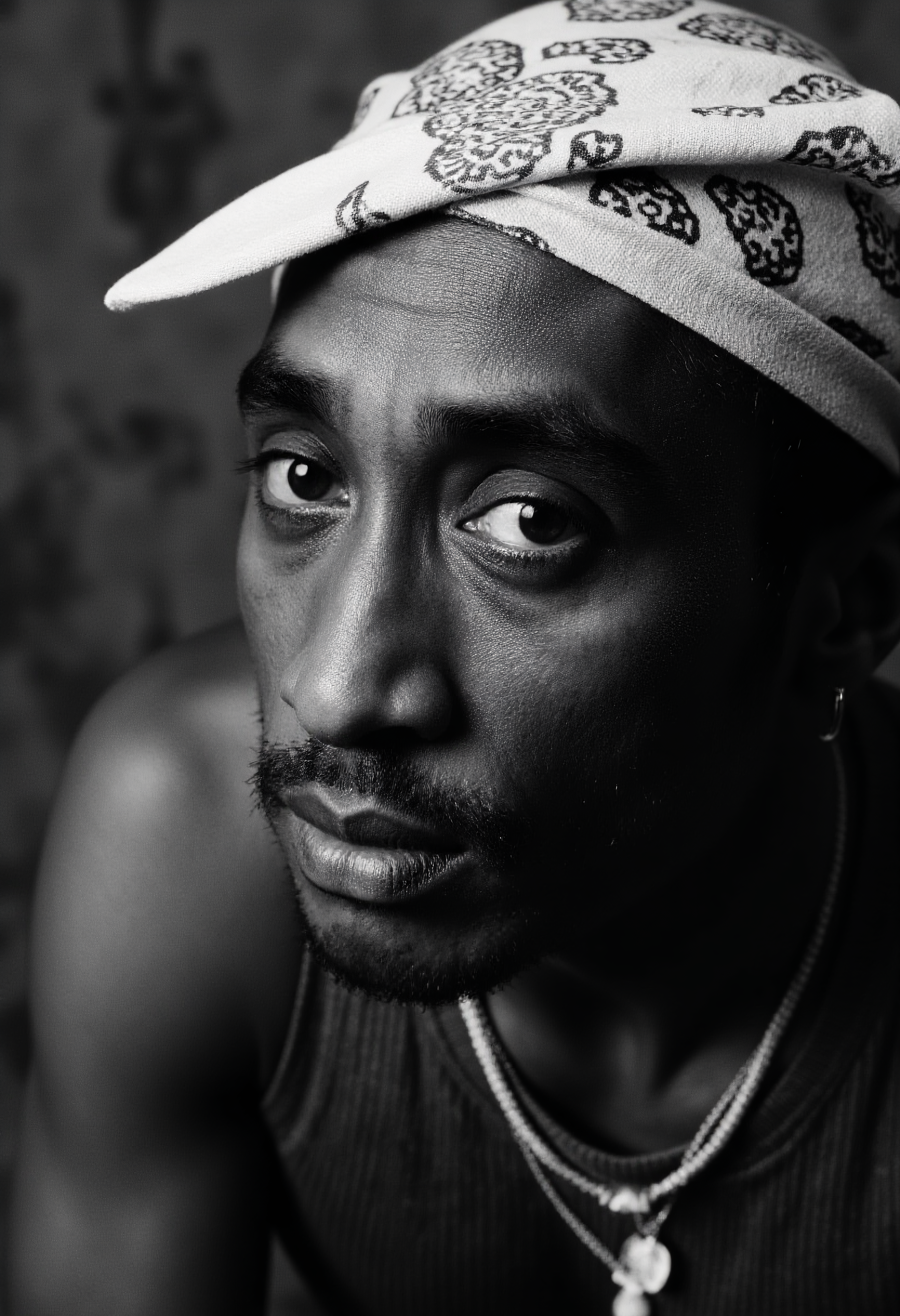 A high-contrast photo of a Tupac Shakur, captured as a black and white portrait, posing for a picture in the style of david-bailey. The subject is centered and gazing directly at the camera, showcasing his expressive eyes and a confident yet thoughtful demeanor. His iconic bandana and minimalistic jewelry enhance the timeless appeal of the image. The background is softly blurred to emphasize the subject, with subtle hints of a gritty urban setting. The lighting accentuates sharp contrasts and detailed textures, creating dramatic shadows that highlight his features. This image is taken with a classic Leica M6 camera, using a standard 50mm lens, capturing the depth and tonal range characteristic of high-end analog photography. <lora:david-bailey-flux1-dev-v1:1>