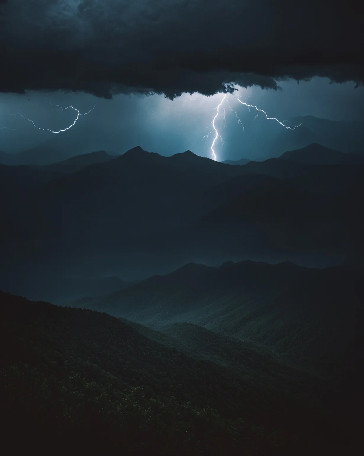 cinematic photography, photo of an endless mountain range, jagged peaks in the dark, no signs of civilization, intense thunderstorm with lightning and pouring rain, storm clouds with visible blue light, full view, Appalachian mountains at night, chiaroscuro lighting, eye level, Canon EOS 5D, Tri-X 400, 50mm, lens flare.