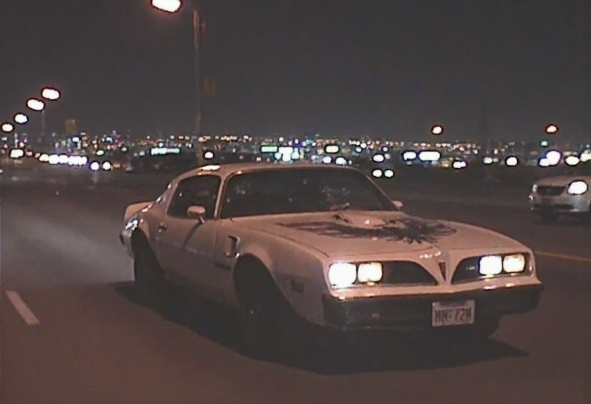 v8s, Dimly lit photo, grungy aesthetic, gritty urban, los angeles city on background, pontiac firebird trans am 1978 driving under high speed, amateur quality, nighttime, in motion, smeared background, headlights glowing, noise and grain film, 