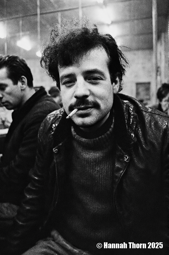 A black and white photograph from a portrait camera angle about a man with a cigarette in his mouth, looking directly at the viewer with a serious expression. the subject, a middle-aged man with curly hair and a mustache, is sitting in the middle of the frame, wearing a black leather jacket over a dark sweater. he appears to be in his late twenties, with a slim body and a slight smile on his face. his eyes are looking straight at the camera, and his hair is styled in a messy manner. he is holding a cigarette between his lips, and there is another man in the background, slightly blurred. the lighting is soft and warm, creating a cozy atmosphere. the background is out of focus, focusing attention on the subject.<lora:_Flux\Anders_Petersen.safetensors:1.0:1.0>