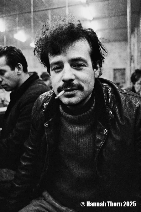 A black and white photograph from a portrait camera angle about a man with a cigarette in his mouth, looking directly at the viewer with a serious expression. the subject, a middle-aged man with curly hair and a mustache, is sitting in the middle of the frame, wearing a black leather jacket over a dark sweater. he appears to be in his late twenties, with a slim body and a slight smile on his face. his eyes are looking straight at the camera, and his hair is styled in a messy manner. he is holding a cigarette between his lips, and there is another man in the background, slightly blurred. the lighting is soft and warm, creating a cozy atmosphere. the background is out of focus, focusing attention on the subject.<lora:_Flux\Anders_Petersen.safetensors:1.0:1.0>