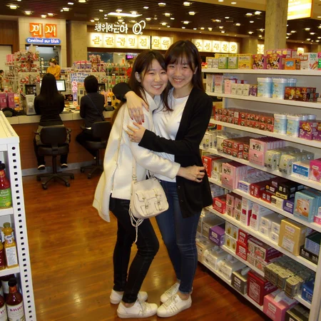 fullbody phone photo of two smiling korean women hugging each other at a busy mall store with a lot of cluttered stuff around them. they are wearing white shoes. flikr photo from 2012. three people working in the background.
