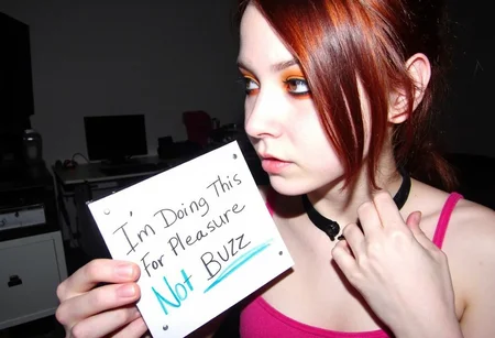amateur photo, low-lit, overexposure, Low-resolution photo, shot on a mobile phone, nighttime, noticeable noise in dark areas. young woman, late teens to early 20s, fair skin, reddish-brown hair pulled back, side-swept bangs covering part of the forehead, dark eyeliner, orange eyeshadow, natural lip color, black collar, pink tank top, relaxed pose, right hand near the neck. The background shows a room with furnitures and desk with computer and monitor. Left hand holding a sign with handwritten text 'I'm Doing This For Pleasure, Not Buzz'. Indoors, low lighting, muted color tones