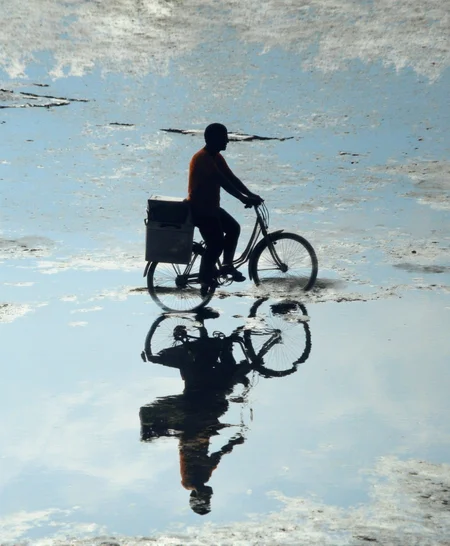 A cinematic photo. A photograph taken from a low angle, looking up at a silhouette of a person riding a bicycle with a box attached to the back, reflected in a puddle on a concrete surface. the image is taken from below, giving a clear view of the shadow of the person riding the bicycle and the reflection of another person in the puddle below. the person on the left is wearing an orange shirt and appears to be in mid-stride, with their arms stretched out in front of them. on the right is the same person, but their reflection is clearly visible. the background is a textured concrete surface with a light blue hue, creating a contrast with the shadows of the two figures. the focus is selectively placed on the cyclist and the person reflected in puddle, making the image stand out.