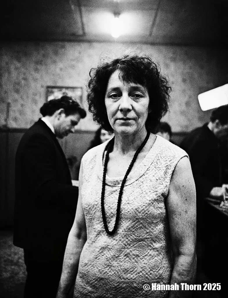 A black and white photograph from a portrait camera angle about a middle-aged woman with curly hair, wearing a knitted dress, standing in a dimly lit room with blurred figures in the background. the woman, who appears to be in her late 60s or early 70s, is standing in the middle of the image, with her upper body facing the viewer and looking directly at them. she has a serious expression and is wearing a sleeveless, lace-like dress with a black beaded necklace around her neck. her curly hair is styled in a way that frames her face and adds to her natural beauty. the background is out of focus, focusing attention on the woman and her outfit.<lora:_Flux\Anders_Petersen.safetensors:0.9:0.9>