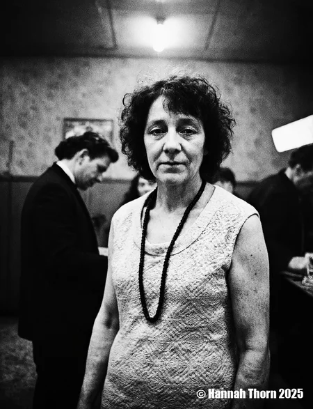 A black and white photograph from a portrait camera angle about a middle-aged woman with curly hair, wearing a knitted dress, standing in a dimly lit room with blurred figures in the background. the woman, who appears to be in her late 60s or early 70s, is standing in the middle of the image, with her upper body facing the viewer and looking directly at them. she has a serious expression and is wearing a sleeveless, lace-like dress with a black beaded necklace around her neck. her curly hair is styled in a way that frames her face and adds to her natural beauty. the background is out of focus, focusing attention on the woman and her outfit.<lora:_Flux\Anders_Petersen.safetensors:0.9:0.9>