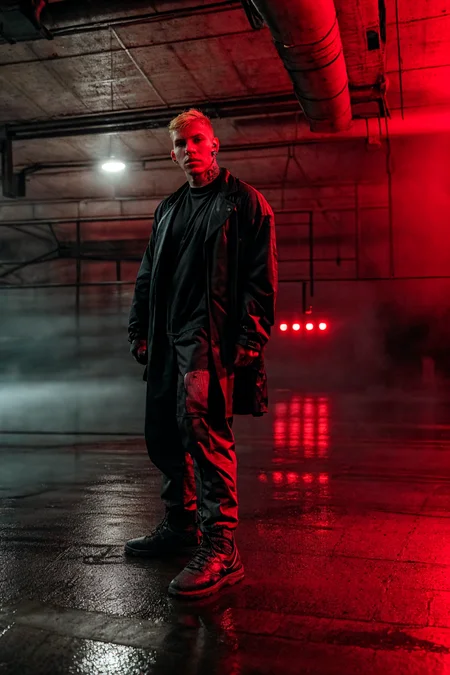 RAW photo, handsome man in dark clothes, techwear, short hair, blonde, tattoos, piercings, muscled, (parking, industrial, wet floor, red lights, volumetric, fog),