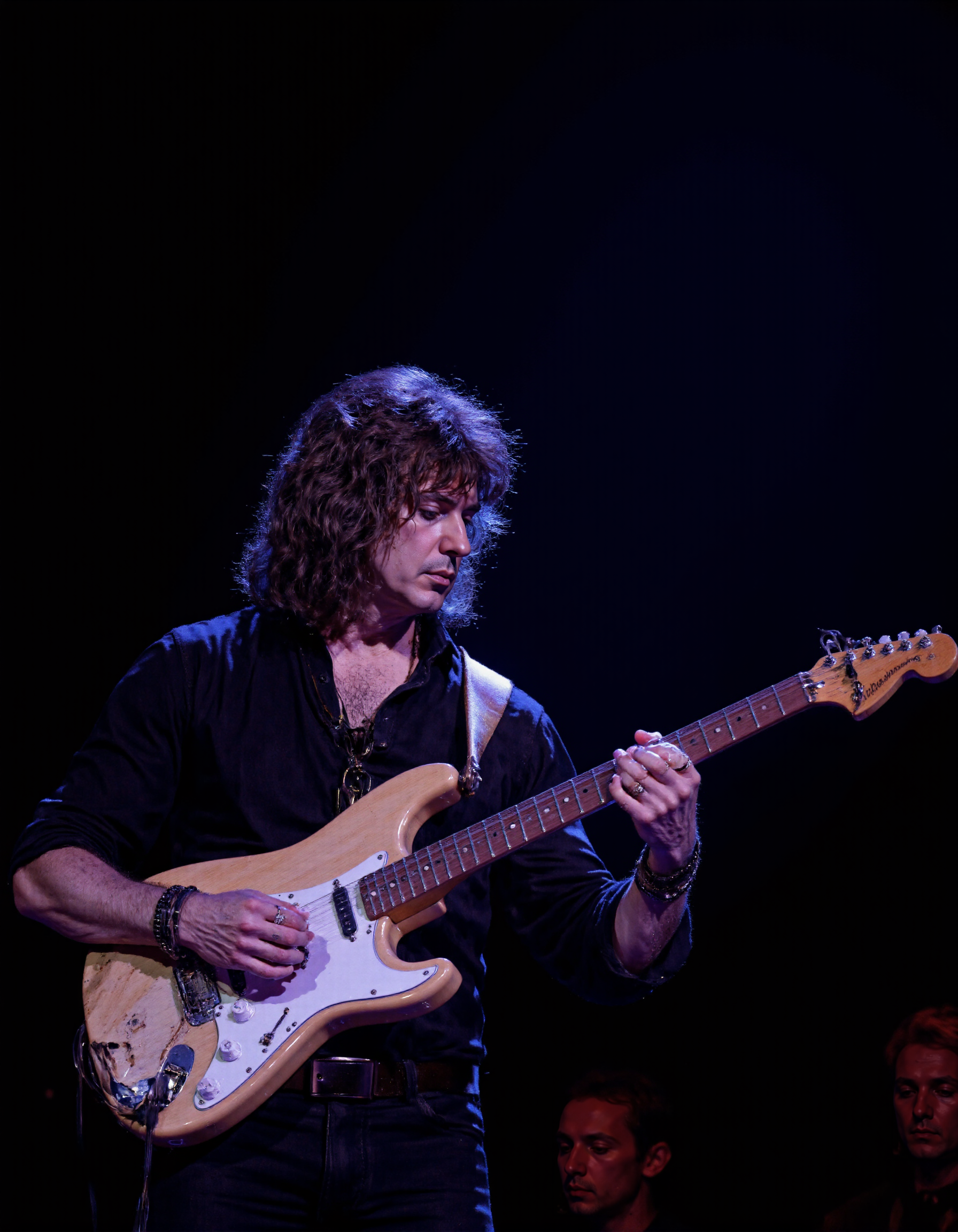 <lora:RICHIE_BLACMORE:1>The image is a photograph capturing a male musician performing on stage. He is centrally positioned, playing an electric guitar. The guitar is a light beige color with white accents, and he is holding it with both hands. The musician has shoulder-length, dark, wavy hair and is wearing a black, long-sleeved shirt. He is also adorned with several bracelets on his right wrist. His facial expression is focused and intense, indicative of deep concentration.
The background is dark, creating a stark contrast that highlights the musician. The lighting is focused on him, casting a soft glow that enhances the texture of his hair and the sheen on the guitar. The overall ambiance suggests an indoor concert setting, possibly a nightclub or small venue, given the intimate lighting and stage setup.
The photograph is a high-resolution, full-color image with sharp details, capturing the textures of the guitar and the musician's clothing. The image emphasizes the musician's connection with his instrument, showcasing the dynamic interplay between performer and instrument. The photograph captures a moment of artistic expression and musical engagement.