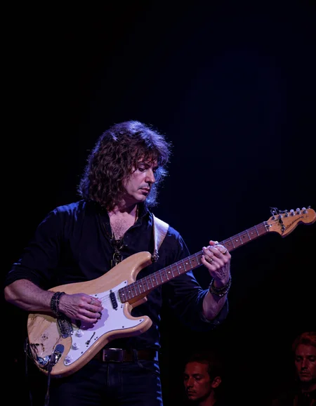 <lora:RICHIE_BLACMORE:1>The image is a photograph capturing a male musician performing on stage. He is centrally positioned, playing an electric guitar. The guitar is a light beige color with white accents, and he is holding it with both hands. The musician has shoulder-length, dark, wavy hair and is wearing a black, long-sleeved shirt. He is also adorned with several bracelets on his right wrist. His facial expression is focused and intense, indicative of deep concentration.
The background is dark, creating a stark contrast that highlights the musician. The lighting is focused on him, casting a soft glow that enhances the texture of his hair and the sheen on the guitar. The overall ambiance suggests an indoor concert setting, possibly a nightclub or small venue, given the intimate lighting and stage setup.
The photograph is a high-resolution, full-color image with sharp details, capturing the textures of the guitar and the musician's clothing. The image emphasizes the musician's connection with his instrument, showcasing the dynamic interplay between performer and instrument. The photograph captures a moment of artistic expression and musical engagement.