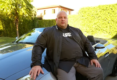photo of an overweight 40 y.o. man sitting in an expensive convertible sports car, sun reflected on paint, wearing suit jacket and t-shirt, bald head combed over, stoic facial expression, sunny day, brightly lit, villa in background <lora:mobile:0.8>