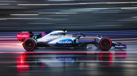 A sensational photograph photograph of an f1 car in motion, with glowing hot brakes. The background and track and wheels are blurred in motion capturing the essence of speed. The race car's brakes are glowing red hot, and are reflected on the wet track. There is a bit of jitter. The lighting is dramatic and creates high contrast, but the image is just a bit washed out due to age. The formula one race car is painted in a delightful gray and blue and magenta scheme with the number 1 and the is sponsored by "NaturalGen". The car is seen in profile, directly from the side. The image is expertly composed and is suitable as an artistic work in minimalist technical photographic style