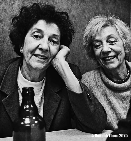 A black and white photograph from a close-up camera angle about two elderly women sitting at a table, smiling and looking at the viewer. on the left side of the image, a woman with curly hair appears to be in her late 60s, wearing a blazer and a white shirt, sitting with her hand on her chin, looking directly at the camera with a neutral expression. she has short, curly hair and a slim body. her eyes are wide open, and she has a slight smile on her face. her hair is styled in a way that frames her face, and her hair color is dark and curly. she is wearing a sweater with a collar, and the woman on the right side is also smiling, showing her teeth. the background is a simple, textured wall, and there is a bottle of beer on the table in front of them.<lora:_Flux\Anders_Petersen.safetensors:0.9:0.9>