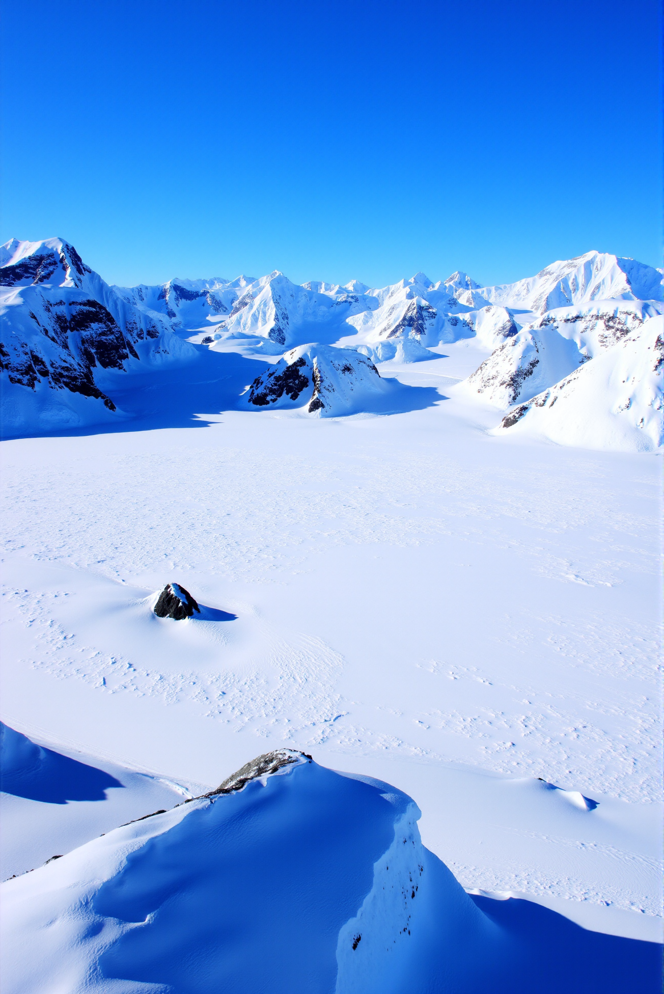 <lora:Ice_Desert_style:0.8> IceDesert style.
Massive glacier fields curve elegantly around steep ridgelines, their surfaces burnished to smooth, silky perfection by relentless wind and ice compression. Towering alpine peaks rise behind, draped in luminous snow that catches the full brilliance of the sky's cobalt blue. Crevasses and ridges form soft, rolling textures across the ice, with subtle shifts in elevation enhancing depth and realism. Captured with a long lens and mid-morning polar light, the image emphasizes clarity and structure through tonal gradation—from deep ultramarine in the shadows to near-white highlights on the upper slopes. Black volcanic rock protrudes in sharp vertical slices from beneath the glacier, adding contrast and visual rhythm. A pure, pristine arctic composition that echoes the tension between permanence and erosion across a tranquil, ice-bound skyline.