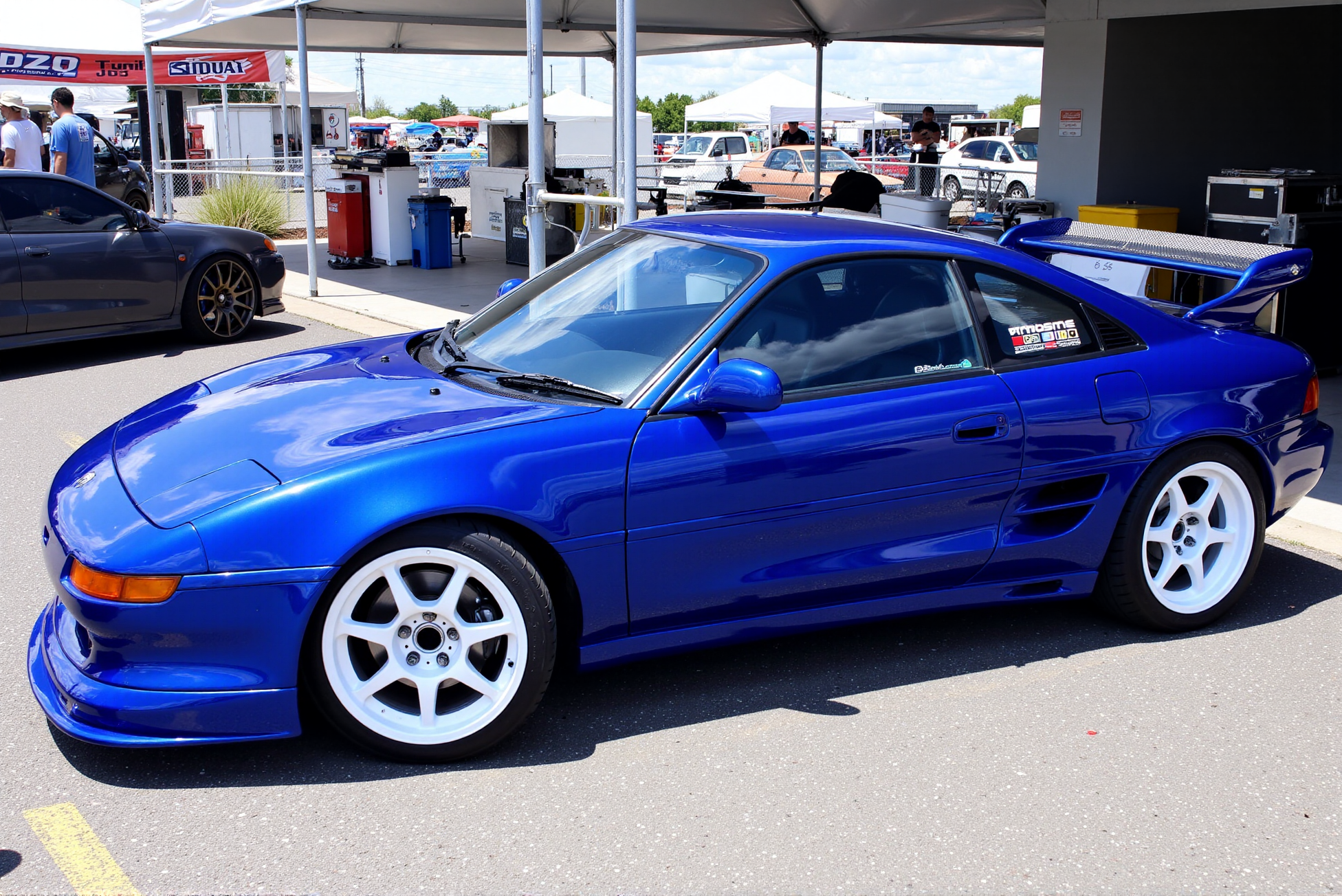 <lora:Toyota_MR2_SW20:0.8> 
A striking blue Toyota MR2 SW20 captured in clean side profile at an outdoor car meet, parked beside tuning booths and performance banners under clear daylight. The deep sapphire paint has a metallic shimmer, with subtle gradients reflecting surrounding vehicles and soft shadow lines. A vented hood, side skirts, and smoothed body kit integrate seamlessly into the coupeâs form. The wheels are white six-spoke racing-style alloys with polished lips, and the stance sits low and even. A large carbon-fiber rear wing provides contrast and aerodynamic balance. Window decals and sponsor markings reinforce the track-built aesthetic. The pavement and background detailing, including toolboxes and race equipment, contribute to the authenticity of the scene. The car exudes confidence and refinement, set in an environment that celebrates performance, individuality, and JDM motorsport culture.