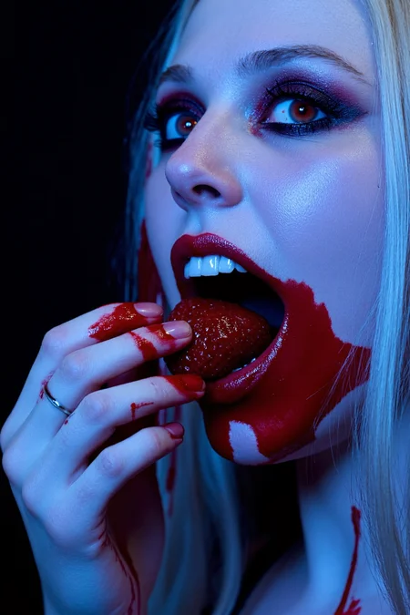 sony_a7III professional style photo, extreme close-up of a young woman in her early 20s, eastern european descent, porcelain skin glowing under dramatic lighting, piercing red eyes locked onto the viewer with an intense, otherworldly gaze, long straight silver hair cascading around her face, dark smoky eyeshadow framing her hypnotic stare, blood-red lips parted to reveal sharp, gleaming fangs, biting deeply into a crimson small strawberry dripping with thick, vivid blood, blood smeared sensually across her lips and fangs for a raw, visceral allure, her glistening tongue teasing through the bloodied bite, seductive expression exuding both danger and allure, side profile subtly accentuating the sharp curve of her jawline, vivid crimson blood streaming from the strawberry, pooling in rich contrast against her pale skin, dark background enveloping her in shadow, high contrast lighting illuminating every detail of her ethereal beauty and the vibrant red of the blood