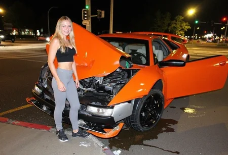 medium-resolution photo, shot on a mobile phone, slightly grainy, with minor visible JPEG artifacts and noise in the darker areas. The image captures an orange sports car lamborghini murcielago heavily damaged in the front, parked askew on a curb at a city intersection. Various angular elements of the damaged hood, express the severity of the impact. The car's right door is open, and there are noticeable wheel marks, debris, and liquid on the road, likely from the impact. Surrounding environment includes a paved sidewalk in the foreground and blurry, partially illuminated street with traffic lights and moving vehicles in the background, street lights casting a yellowish glare, suggest night time in an urban setting, dynamics of urban accidents, car culture. on foreground in front of the car is standing young woman and smile, dynamic pose, she is blonde with long hair, wear grey leggings and black crop top and sneakers