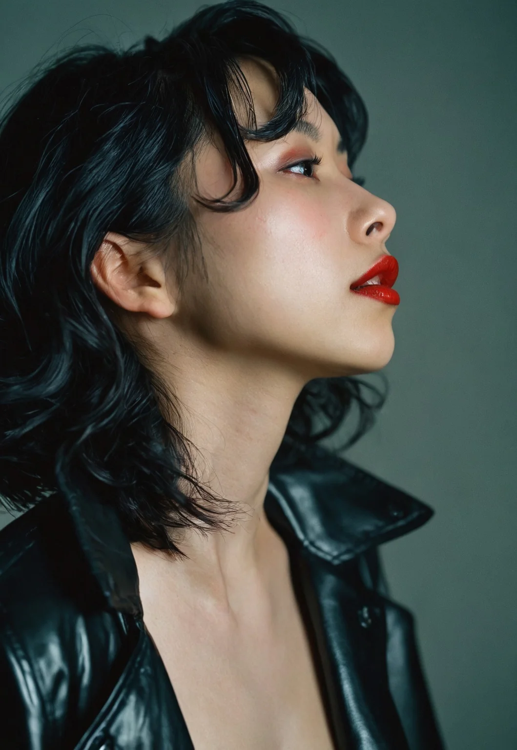 shot on Nikon F3, Close-up portrait, 28 years old badass looking slim japanese girl, side view, sharp jawline, black wavy short hair, pale glowing skin, deep red lipstick, wearing black leather jacket, soft highlights, soft natural lighting, dim dark-ish background, Superia Xtra 400 Film, film emulation, 35mm film, grainy, fIlm grain, halation, Bloom, high dynamic range, film photography, 23mm focal length, iso 100, f/2.8, (sharp details, high quality, 4k, intricate details, highly-detailed)