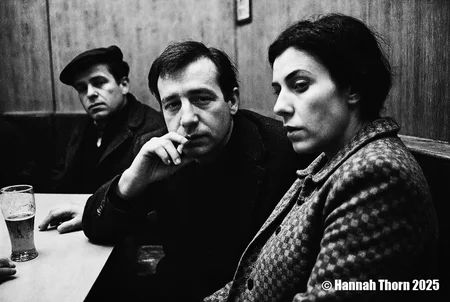 A black and white photograph from the side, featuring two people sitting at a table in a dimly lit restaurant. the man, on the left, is leaning over the table, holding a cigarette in his hand, while the woman, sitting on the right, is facing the viewer with a serious expression. the woman has short, dark hair and is wearing a tweed jacket with a checkered pattern. on the table in front of them, a glass of beer is visible. in the background, a man in a hat and coat is blurred, adding to the moody atmosphere of the scene.<lora:_Flux\Anders_Petersen.safetensors:0.9:0.9>
