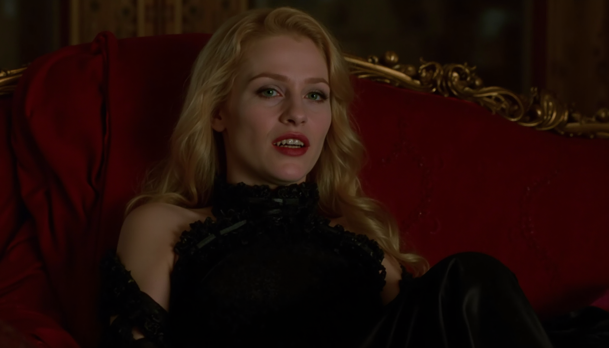 cinematic medium shot of an aristocratic, deathly pale young vampire woman with fangs in an opulent victorian black silk gown, high neckline, reclining on a red velvet couch at night, warm candle light. She has long blonde hair and is looking at the viewer with a haughty, bored expression, slightly open mouth with fangs, black makeup around eyes. Taken from a high angle, analogue film, IMAX, photorealistic, very detailed <lora:iwtv_flux_1_0:1>
