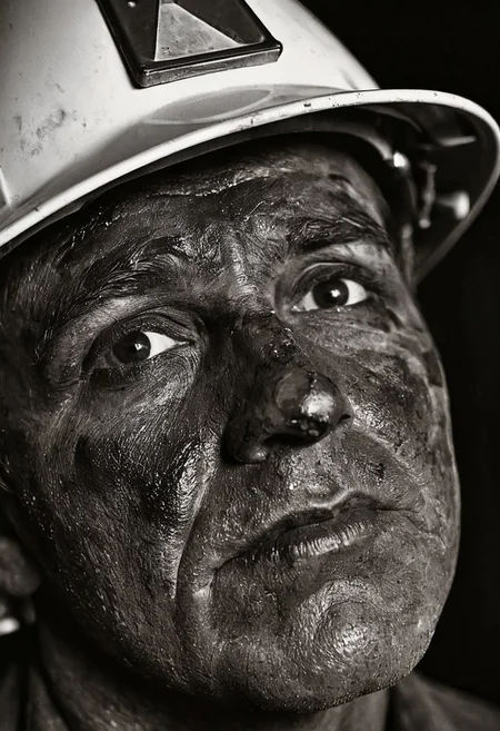 A close-up portrait photograph of a coal miner's face, focusing on his eyes beneath a coal-stained brow. His face is covered in black coal dust, creating stark contrast against the whites of his eyes. The miner wears a traditional hard hat with a headlamp, and his weathered skin shows traces of coal residue. The background is dark and out of focus, suggesting the underground mining environment. Black and white documentary photography with dramatic side lighting and sharp focus on facial features. 4k, intricate details, highly detailed, hyper-realistic, 8k RAW Editorial Photo. cinematic light, dramatic light, shallow depth of field, high budget, cinemascope film grain ISO 200 faded film, 35mm photo, grainy, vignette, vintage, Kodachrome,