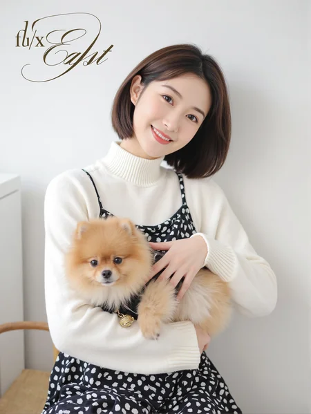 This is a clear and high-resolution everyday photo with a minimalist and natural style. In the picture, there is a woman wearing a white high-neck sweater and a black sundress with white polka dots. She has shoulder-length hair and is smiling as she looks at the camera. In her arms, she is holding a brown Pomeranian dog, which is dressed in a black and white outfit and wears a small golden bell around its neck. The background is an indoor setting with white walls and faint outlines of furniture.
There is a prominent artistic font above the picture, which reads' fluxEast 'in English