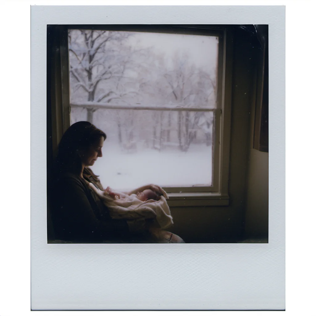 polaroid of A mother, cradling a newborn, looking out the window as the first snow of winter falls gently. <lora:Polaroid_XL:1>