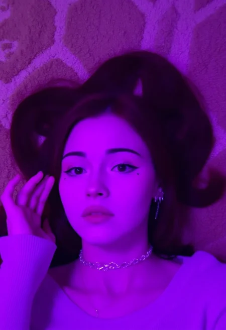 The image is a photograph of a young woman KTNK with a fair complexion and long, straight, dark brown hair styled in two pigtails. She has a petite build and is lying down, possibly on a textured, plush surface with a geometric pattern in shades of beige and brown. The lighting in the image is predominantly purple, casting a cool, ethereal glow over her features.