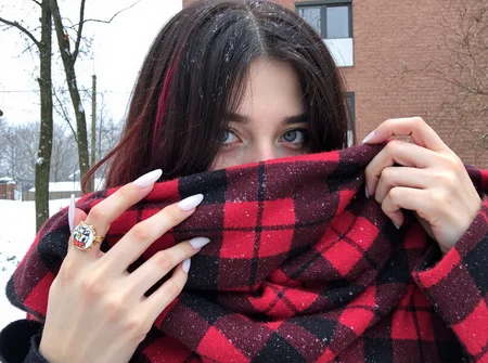 s2ms8ng. Close-up photo of a young woman, mid-20s, partially covering her face with a large plaid scarf, holding the scarf with her hand, revealing long white manicured nails adorned with a cartoon character ring. She has straight, shoulder-length dark hair with red highlights, barely visible due to the scarf. Light snowflakes rest on her hair, suggesting a cold, snowy day. Blue-gray eyes are prominent, her expression hidden by the scarf, creating a mysterious effect. She is outdoors, urban background with a brick wall and bare trees, winter season is evident, her face very underexposed and shadowed
