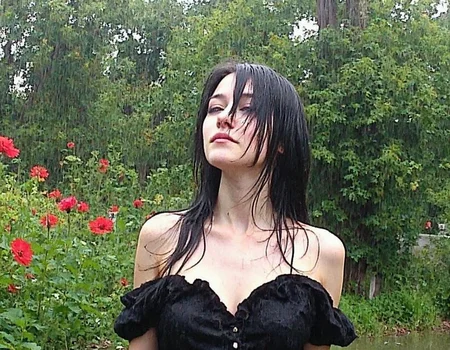 v8s, Young woman looking upwards, fair-skinned, wet hair clinging to face, closed eyes, long straight black hair, wearing black wet dress with ruffled collar, standing amidst lush greenery, vibrant red flowers surrounding, heavy rain falling, outdoor garden setting, overcast lighting, raindrops visible in mid-air, close-up angle, natural lighting conditions