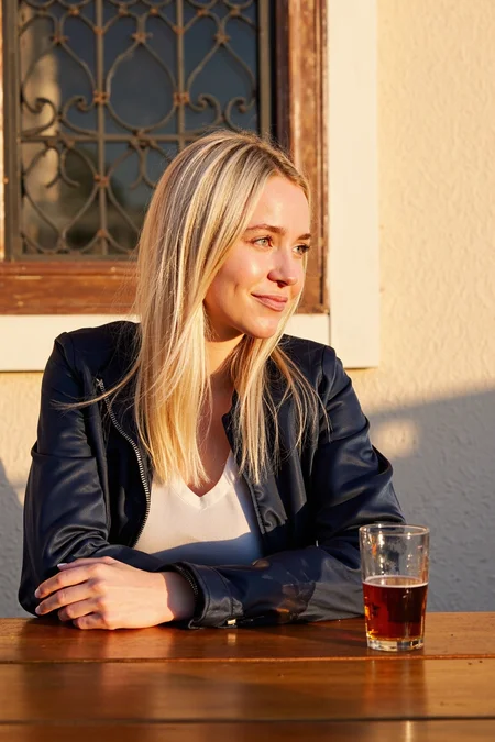 sony_a7III professional style photo, Female, early 20s, fair-skinned, slim build, long straight blonde hair neatly framing face, dark navy jacket partially zipped revealing light-colored inner layer, clear glass of amber beer with slight foam placed on a polished wooden surface, outdoor setting with rustic iron latticework window visible in the background, textured wall surface adding character to the environment, soft warm lighting casting a golden glow on the subject, subtle shadows creating depth, slight smile conveying relaxed expression, side gaze directed away from the camera, medium camera angle capturing upper body, warm ambient evening lighting enhancing natural tones, serene and inviting atmosphere, casual yet refined outdoor scene