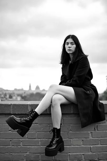 sony_a7III professional style photo, Black and white photo of a young woman sitting on a brick wall, legs extended out in front of her, black platform combat boots prominently in the foreground. She has light skin, long black hair, and is wearing a dark trench coat. She has a slender build and a relaxed, introspective expression. The background shows a cloudy sky and a distant cityscape. The lighting is natural, with soft shadows. The camera angle is low, looking up at her. shallow depth of field, background slightly blurred