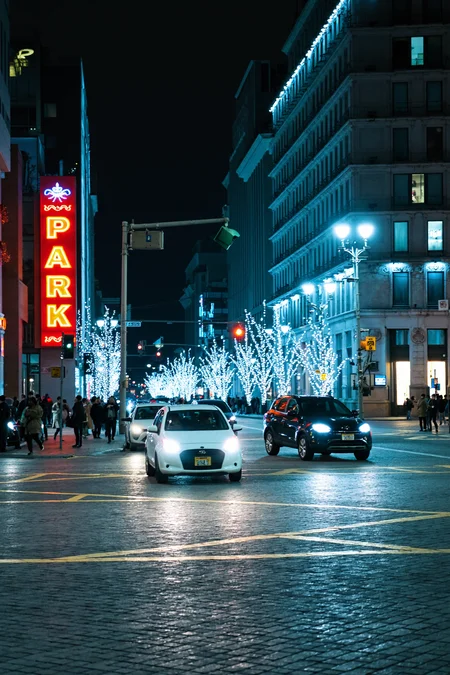 sony_a7III professional style photo, Nighttime city street bustling with cars, white compact sedan and dark SUV in motion alongside other indistinct vehicles, glowing streetlights casting a soft warm glow, vibrant red and orange illuminated PARK signage on the left, buildings adorned with decorative string lights creating a festive atmosphere, scattered pedestrians on sidewalks engaged in evening activities, brightly lit street intersection with visible traffic signals, yellow dividing lines cutting through uneven cobblestone road surface, reflections from bright car headlights adding dynamic light patterns, urban environment featuring tall buildings with a blend of modern glass exteriors and classic brick facades, faintly visible storefronts in the distance, clear night sky with no visible stars, cool color palette dominated by blues and greys accented by warm streetlight hues, cinematic depth of field enhancing foreground clarity, slight motion blur on moving vehicles, low-angle perspective emphasizing the grandeur of the buildings, artificial lighting creating vivid contrasts, nighttime cityscape setting