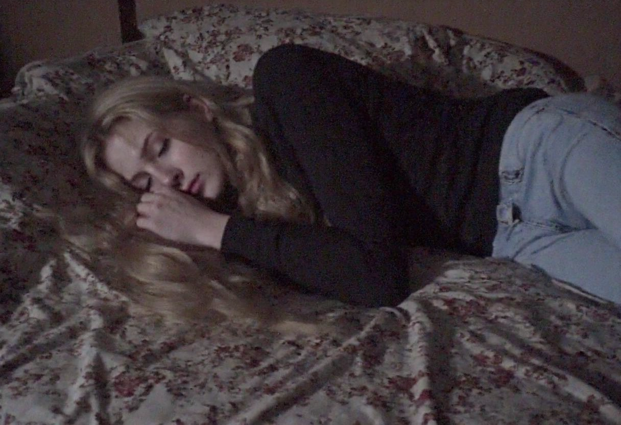 v8s, Blonde-haired woman, lying on the bed, sleeping, closed eyes, long wavy hair, dark long-sleeve top, light blue jeans, indoor setting, detailed patterned bedspread, muted dark colors, dim lighting, patterned fabric with floral design, cozy and intimate atmosphere, underexposed image. Amateur quality, Analog distortion, low resolution, Chromatic aberration, tape noise, Grainy texture, Frame jitter, muted colors, overexposed foreground