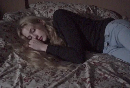 v8s, Blonde-haired woman, lying on the bed, sleeping, closed eyes, long wavy hair, dark long-sleeve top, light blue jeans, indoor setting, detailed patterned bedspread, muted dark colors, dim lighting, patterned fabric with floral design, cozy and intimate atmosphere, underexposed image. Amateur quality, Analog distortion, low resolution, Chromatic aberration, tape noise, Grainy texture, Frame jitter, muted colors, overexposed foreground