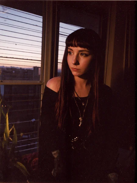 p0l2rd, amateur photo bedroom, window, sunset, low light, blinds, plants, quilt, patchwork quilt, cozy, interior, evening, calm atmosphere, urban skyline, silhouette, warm colors, twilight, soft lighting, home decor, tranquil, intimate setting, goth teen girl, gothic fashion, off-shoulder top, corset, dreadlocks, eccentric hairstyle, dark makeup, facial piercing, gloves, alternative fashion, smirk, edgy style. prominent film grain, high contrast lighting casting dramatic shadows, grainy film-like texture, overexposed and blurry photo