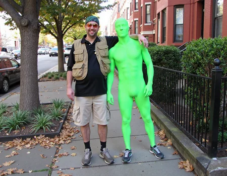 A full-body shot of two men standing side by side on a city sidewalk, posing for the camera. On the left, the first man is dressed casually in an outdoor or adventurer-style outfit. He is wearing a black polo shirt, a beige utility vest with multiple pockets, beige cargo shorts, and hiking boots. He accessorizes with a green bandana tied around his head and orange-tinted sunglasses. He has a cheerful expression and is smiling broadly, showing his teeth. His right arm is draped around the shoulders of the man standing next to him, in a friendly and relaxed posture. The second man is wearing a bright, neon green full-body spandex suit that completely covers his body, including his face, creating a smooth, featureless appearance. The suit hugs his form tightly, and he is wearing running shoes, adding a quirky and playful element to his outfit. His body language is neutral, and due to the spandex covering his face, no facial expression is visible. In the background, there is a row of red brick buildings, typical of an urban residential area, with neatly manicured bushes and trees. The scene suggests a casual, lighthearted moment between friends or participants in an event, with the costumes adding a sense of humor and fun to the shot. The sidewalk and surrounding foliage indicate it is an autumn day, with a few fallen leaves visible on the ground <lora:amateurphoto-v6-forcu:0.8>