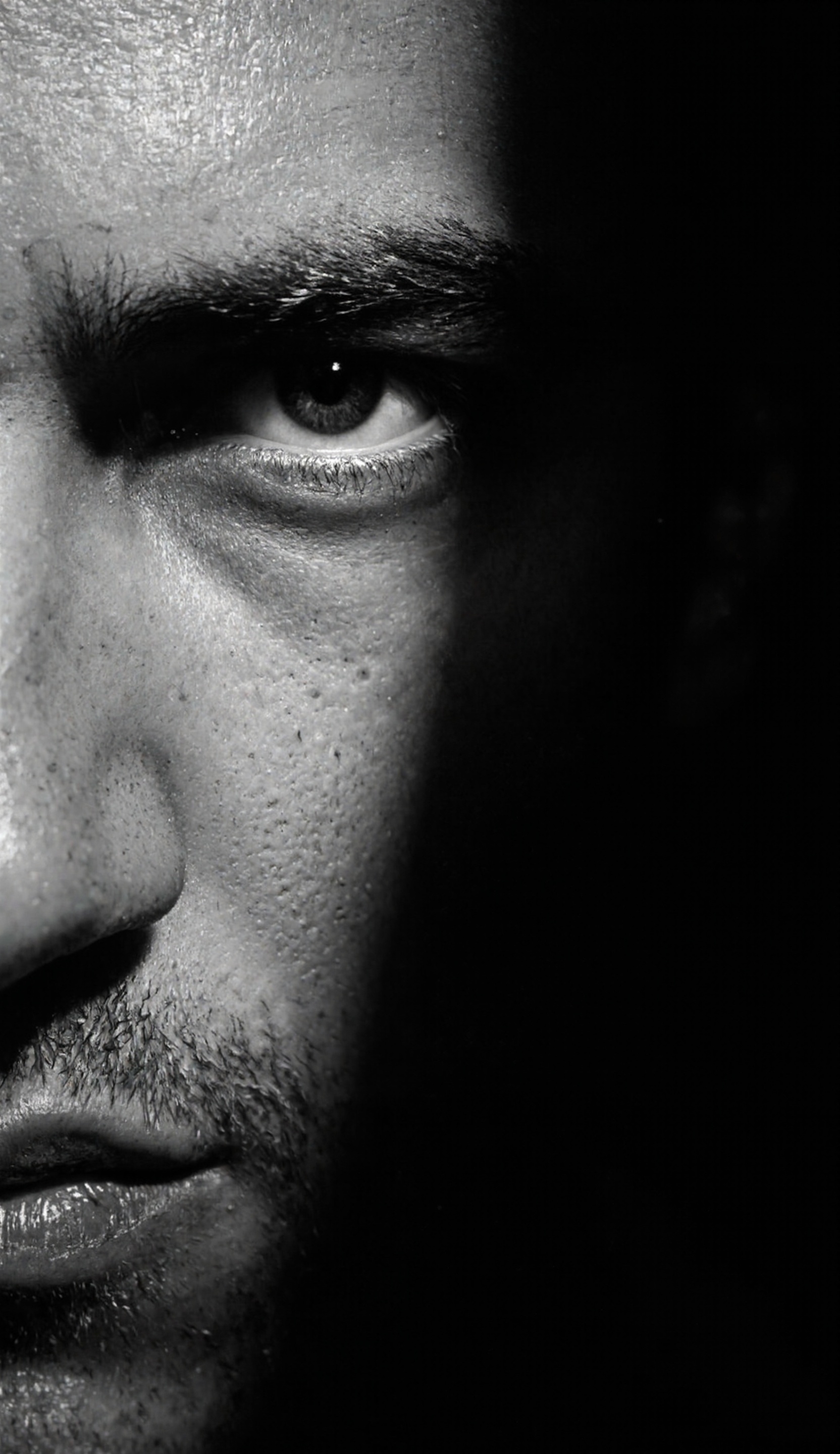 A dramatic, high-contrast black and white portrait photograph, reminiscent of film noir, focuses on a man’s face. Strong side lighting casts deep shadows across one side of his face, emphasizing the angularity of his cheekbones and jawline. The light source appears to be positioned high and to the left, creating a stark division between light and shadow. 
His expression is intense and brooding, with a slight furrow in his brow and a direct, unwavering gaze. The man has short, dark hair, neatly styled, and visible stubble suggesting a rugged, masculine appearance. His eyes are dark and piercing, conveying a sense of determination or perhaps melancholy. 
The texture of his skin is visible, with subtle imperfections and lines adding to the realism of the portrait. The background is completely black, isolating the man’s face and intensifying the dramatic effect. The composition is tightly cropped, focusing solely on his head and shoulders. 
The overall mood is one of mystery, intensity, and perhaps a hint of danger. The high contrast and dramatic lighting create a sense of tension and intrigue. The photograph appears to be a studio portrait, carefully composed and lit to emphasize the man’s features and convey a specific emotional state. The style evokes classic Hollywood portraits of the 1940s and 1950s, with a focus on strong lighting and dramatic composition.