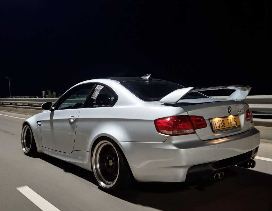 s2ms8ng, visible sensor noise, artificial over-sharpening, heavy HDR glow. rear three-quarter view of a silver sports car bmw m3. The car appears to be driving on high speed on a dimly lit highway at night. car with custom body parts. The background reveals a guardrail and a dark, empty road, intensifying the isolation of the scene, low lighting, in motion, smeared details
