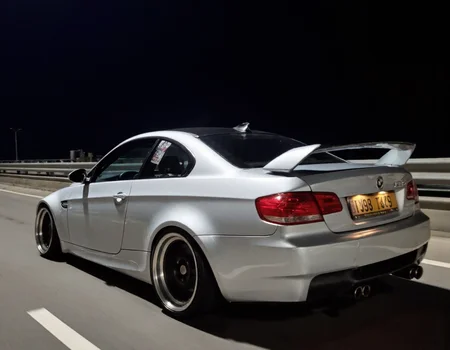 s2ms8ng, visible sensor noise, artificial over-sharpening, heavy HDR glow. rear three-quarter view of a silver sports car bmw m3. The car appears to be driving on high speed on a dimly lit highway at night. car with custom body parts. The background reveals a guardrail and a dark, empty road, intensifying the isolation of the scene, low lighting, in motion, smeared details