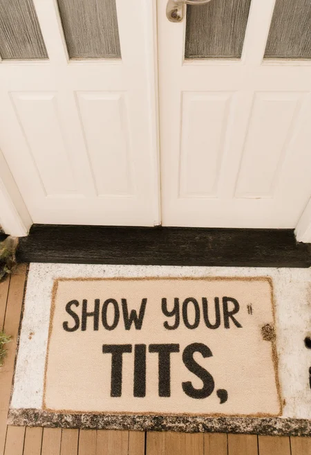 This photograph captures a cozy and welcoming entrance scene. The image features a small, rectangular coir doormat placed in front of a white door with the text "Show Your Tits" written in all caps.