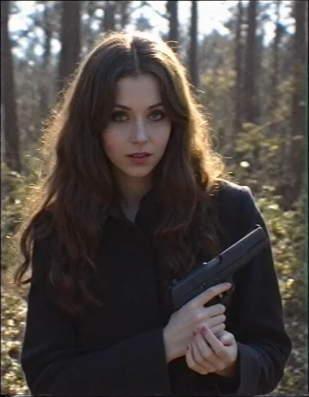 v8s, Young woman, early 20s, casual style, holding pistol, wearing dark trench coat, long wavy brown hair cascading over her shoulders, looking direct at the viewer, gothic heavy makeup, outdoors in the dark forest, autumn, sunlight filtering through the trees. Amateur quality, Analog distortion, low resolution, Chromatic aberration, tape noise, Grainy texture, Frame jitter, muted colors, overexposed foreground