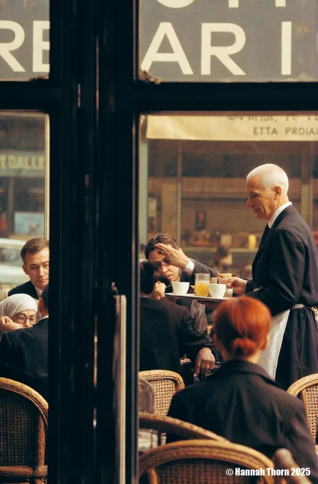 The image captures a scene within a Parisian café, viewed through a darkened vertical element, likely a door frame or window mullion. The focus is on a waiter, an older man with close-cropped white hair, dressed in a dark suit jacket and white apron, holding a tray laden with drinks and cups. He appears to be serving a group of patrons seated at wicker chairs. The patrons are less distinct, partially obscured by the foreground element and the soft focus of the image. A woman with a light-colored head covering and glasses is visible in the lower left corner, seemingly absorbed in something she holds in her hands. Other figures are present, including a man with dark hair who is partially obscured by the frame, and a woman with reddish hair who appears to be raising her hand to her face. The overall style of the image evokes a candid, street photography aesthetic. The muted color palette, dominated by browns, grays, and dark tones, contributes to a vintage or nostalgic feel. The soft focus and grainy texture, along with the slightly blurred background, suggest that the photo might have been taken with a slower shutter speed, perhaps capturing a fleeting moment in the café's life. The composition, with the dark vertical bar in the foreground partially obscuring the scene, creates a sense of depth and voyeurism, as if the viewer is catching a glimpse of a private moment. The blurred background, including what appears to be signage or lettering on the wall behind the waiter, adds to the impression of a bustling, lively atmosphere. The lighting is diffuse, likely natural light filtering into the café, contributing to the overall ambiance of the scene. The style could be described as realistic, capturing the everyday life of a Parisian café in a naturalistic manner. <lora:_Flux\Saul_Leiter_Streetphotography.safetensors:1.0:1.0>