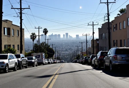 This appears to be the same Los Angeles street scene I described earlier: Urban residential street view, downtown Los Angeles skyline visible in background through morning haze, steep hill street perspective, parked cars lining both sides of road, utility poles with multiple power lines crossing image, mid-century apartment buildings visible on left side, mixture of vehicles including postal truck, SUVs, sedans, palm tree visible in middle distance, beige-colored residential buildings, typical LA neighborhood architecture, morning sunlight creating atmospheric haze effect, clear blue sky above skyline, downtown skyscrapers appearing silvery-white in distance, street markings visible on asphalt, parallel parking arrangement along curbs, moderate telephoto compression effect on cityscape, early morning lighting conditions, slight lens flare visible, urban residential atmosphere