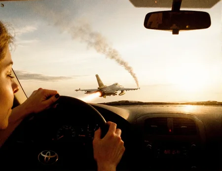 8n8log,View from the driver’s seat of a Toyota car flying high above the clouds, young woman's hands gripping the steering wheel tightly, she has black nailpolish, dashboard gauges illuminated. Through the windshield, a fighter jet f16 is visible from the back, flying away into the distance, its twin afterburners glowing brightly with flames and leaving long, smoky vapor trails across the bright, cloud-filled sky. The sun is low on the horizon, casting warm light and subtle lens flares across the windshield. The scene captures a surreal, dynamic moment with the jet's engines prominently displayed, emphasizing its motion and power. high contrast, film photography aesthetic, film grain effect prominent throughout image, high contrast lighting creating dramatic shadows, grainy film-like texture