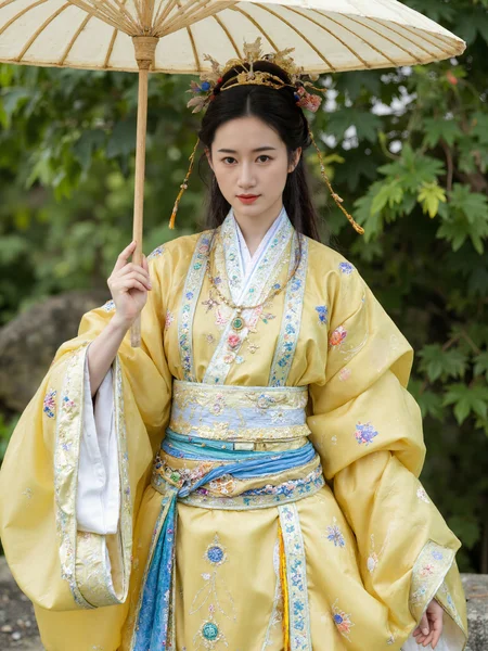 This is a high-resolution photograph featuring a young Asian woman in traditional Chinese attire. She stands in a natural outdoor setting, surrounded by blurred greenery, suggesting a park or garden. The woman, with fair skin and delicate features, wears an elaborate, flowing yellow and blue silk robe adorned with intricate embroidery and gold accents. Her hair is styled in an elegant updo, with ornate hairpieces and jade accessories. She holds a large, traditional Chinese parasol above her head, adding a sense of grace and historical authenticity. The lighting is soft and natural, enhancing the serene and picturesque ambiance. The camera angle is at eye level, capturing her full figure and the detailed textures of her clothing. The composition follows the rule of thirds, with the woman positioned slightly off-center, drawing the viewer's eye naturally across the image.