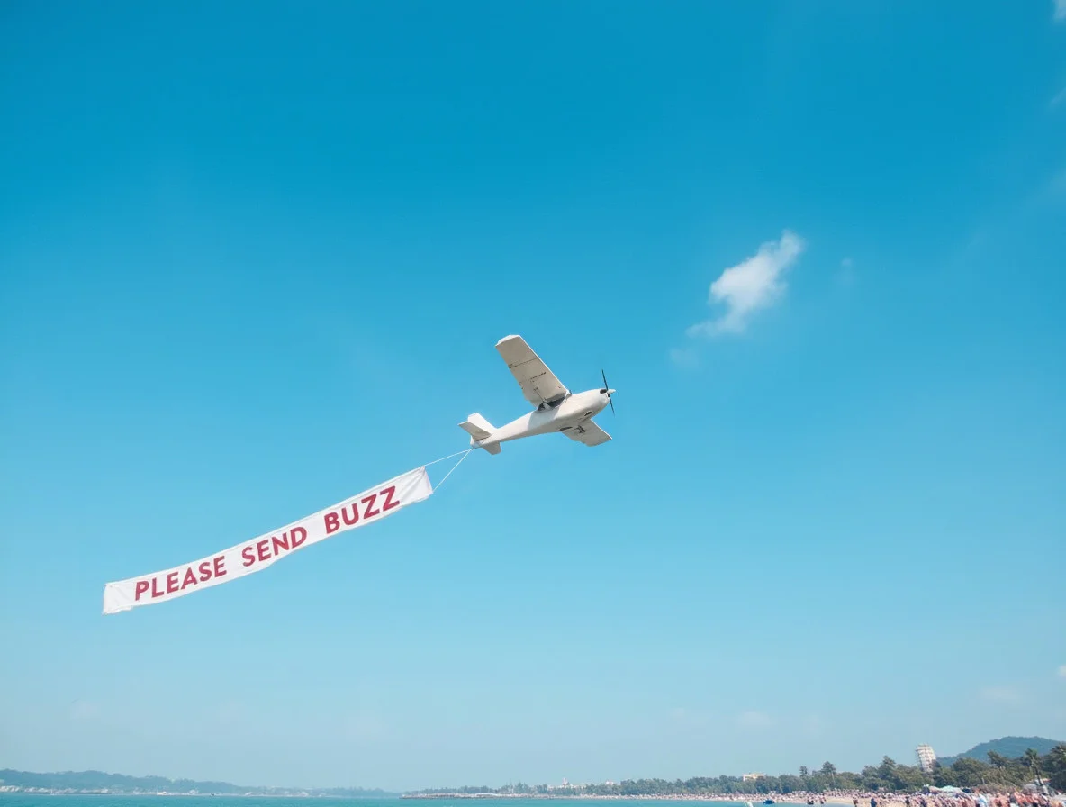 small white airplane towing a long banner that reads "PLEASE SEND BUZZ" across a vibrant, clear blue sky, flying over a crowded beach