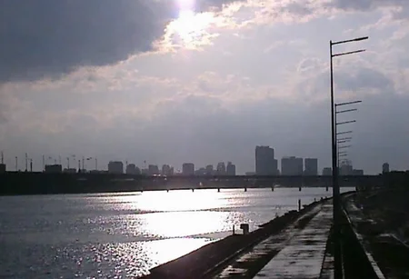 v8s, Overcast sky with dense clouds, sun peeking through, strong reflections on water, wide river or body of water, distant bridge spanning water, modern city skyline silhouette, high-rise buildings in background, empty road alongside water, tall streetlights lining road, wet pavement suggesting recent rain, early morning or late afternoon light, hazy atmosphere, dim lighting, outdoor setting, high contrast between bright reflections and dark clouds